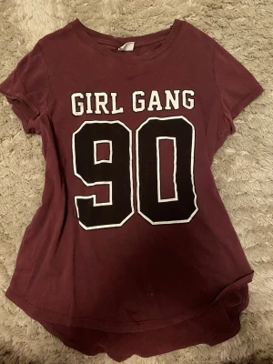 Vinröd t-shirt med tryck GIRL GANG 90 - Snygg vinröd t-shirt med stort vitt och svart tryck på framsidan där det står 'GIRL GANG 90'. T-shirten har rund halsringning och korta ärmar. Perfekt för en avslappnad och cool stil. Små hål som du see på bild tre med lätt att sy ihop själv med nål och tråd 