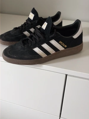 Adidas Spezial svarta sneakers mocka - Säljer ett par klassiska Adidas Spezial sneakers i svart mocka med vita detaljer och bruna gummisulor. Skorna har de ikoniska tre ränderna på sidan och guldfärgad SPEZIAL-text. Perfekta för dig som gillar retrostil och streetwear.