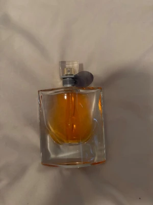Elegant parfymflaska med orange doft - Säljer en stilren parfym i en fyrkantig glasflaska med transparent lock och silverfärgad spraydel. Vätskan är orange och flaskan har en modern, elegant design som passar perfekt på sminkbordet.