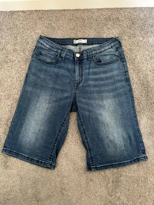 Blå denimshorts med fem fickor - Säljer ett par klassiska blå jeansshorts i denim med fem fickor och knappgylf. Modellen har normal passform och längden går till knäna. Perfekta för varma dagar och enkla att matcha med olika stilar.