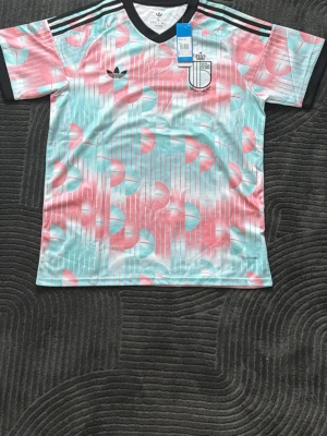 Adidas retro fotbollströja XL - Säljer en färgstark retroinspirerad fotbollströja från Adidas i storlek XL. Tröjan har ett unikt mönster i ljusblått, rosa och vitt med svarta detaljer och klassiska Adidas-ränder på axlarna. Perfekt för dig som vill sticka ut på planen eller gatan.