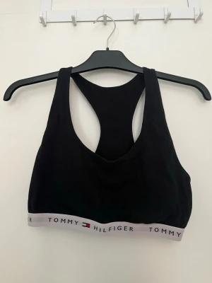 Svart bh från Tommy Hilfiger - Svart sport-bh från Tommy Hilfiger med brottarrygg och bred resår nertill där loggan syns i vitt, rött och blått. Tillverkad i mjukt och stretchigt material för en bekväm passform. Strl M/L.