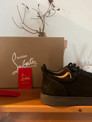 Svarta sneakers från Christian Louboutin - Stilrena svarta sneakers från Christian Louboutin i mocka med ton-i-ton snörning och diskret logga på sidan. Skorna har en klassisk låg siluett och svart sula. Perfekta för dig som gillar exklusiva och minimalistiska sneakers. Om mitt pris inte passar, buda på. Bjuder på en tvättning innan jag fraktar. Dom är 27,5 cm långa. Skambud undanbes! MVH 