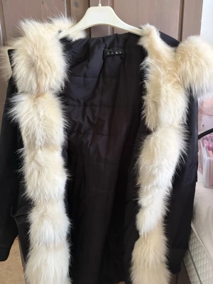 Svart parkas med äkta kanin päls - Snygg svart parkas med stor luva och fluffiga kanin päls i vitt längs huvan och öppningen. Jackan har en rak passform och är vadderad för extra värme. Perfekt för kalla dagar när du vill hålla stilen.