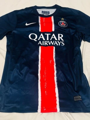 PSG matchtröja blå röd Nike - Snygg PSG matchtröja i mörkblått med en bred röd och vit rand i mitten. Framsidan har Qatar Airways-logga och PSG-emblem med stjärna. Baksidan har D.Doué och nummer 14 i rött och vitt. Tillverkad i lätt och ventilerande material, perfekt för fotboll eller träning.