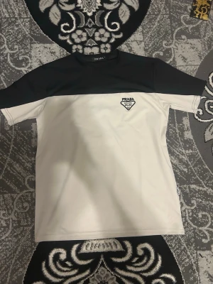 Svartvit t-shirt från Prada - Snygg svartvit t-shirt från Prada med broderad logga på bröstet. T-shirten har svart överdel och vita partier nedtill och på ärmarna. Klassisk passform och rund halsringning. Perfekt för dig som gillar stilrena designer.