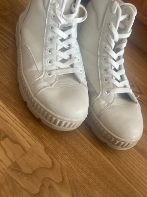 Vita höga sneakers med platåsula - Säljer ett par vita höga sneakers med kraftig platåsula och vita skosnören. Skorna har en stilren design med paneler i både slätt och mockaliknande material. Perfekta för dig som vill ha en clean och trendig look.