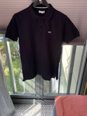 Svart pikétröja från Lacoste - Klassisk svart pikétröja från Lacoste med korta ärmar och krage. Tröjan har knappar framtill och den ikoniska krokodilloggan på bröstet. Perfekt till jeans eller shorts för en clean och stilren look.