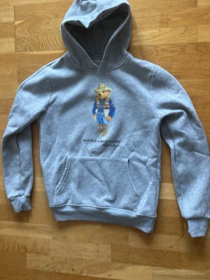 Grå hoodie med Polo Bear-tryck - Säljer en grå hoodie från Ralph Lauren med det ikoniska Polo Bear-trycket på bröstet. Hoodien har huva, magficka och ribbade muddar vid ärmslut och nederkant. Perfekt för en avslappnad och trendig look.
