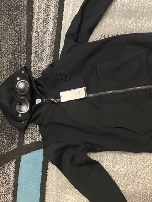 Svart hoodie med glasögon från C.P. Company - Säljer en svart hoodie från C.P. Company med dragkedja och ikoniska glasögon i huvan. Hoodien har två stora fickor framtill och diskret broderad logga på bröstet. Perfekt för dig som vill sticka ut med en unik streetwear-look.