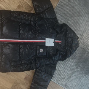 Svart Moncler Jacka - Snygg svart pufferjacka med huva och dragkedja framtill. Jackan har en röd, vit och blå rand längs dragkedjan och en logga på bröstet. Perfekt för kalla dagar och har praktiska fickor med dragkedja.