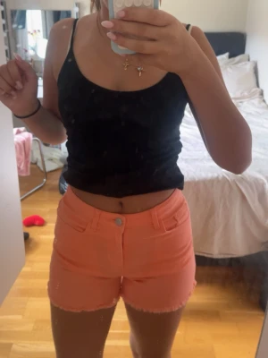 Korallfärgade jeansshorts midwaist - Säljer ett par korallfärgade jeansshorts med rå kant nertill och klassisk femficksdesign. Shortsen har midwaist passform och stängs med dragkedja och knapp. Perfekta för varma dagar och ger en färgklick till din outfit.