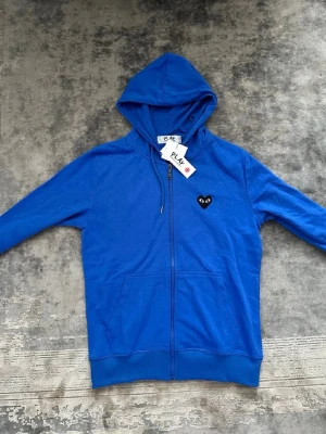 Blå hoodie med svart hjärtdetalj - Säljer en blå hoodie med dragkedja och huva. På bröstet finns ett svart hjärta med ögon och på ryggen ett större hjärta i samma stil. Hoodien har ribbade muddar och fickor framtill. Perfekt för dig som gillar streetwear och vill sticka ut.