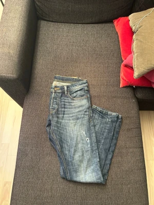 J&J Slim Glenn  - Ett par blå jeans med slim fit och slitningar på låret. Jeansen har klassisk femficksdesign, dragkedjegylf och kontrastsömmar. Materialet är denim i bomull och färgen har en tvättad look med ljusare partier. Storlek=w29, l30