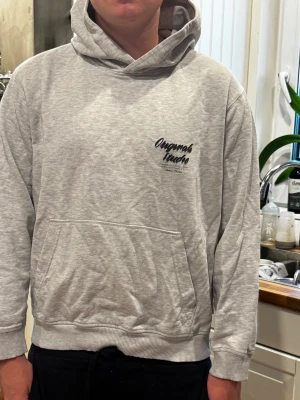 Ljusgrå hoodie Originals Studio - Ljusgrå hoodie med tryck 'Originals Studio' i svart på ryggen och mindre logga på bröstet. framtill och ribbade muddar. Skön och avslappnad passform, perfekt för chill dagar.