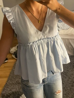 Ljusblå volangblus med v-ringning - Supersöt ljusblå topp med volangärmar och v-ringning fram. Modellen är lös och luftig med rynk under bysten och knytning i ryggen. Perfekt till jeans eller kjol för en fräsch look. 