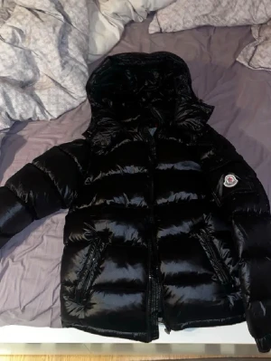 Svart dunjacka från Moncler - Helt ny snygg och varm Moncler Maya i svart – en riktig vinterfavorit. Jackan är fylld med vitt ankdun (ca 90 % dun), vilket ger utmärkt volym och värme under kalla dagar.   Detaljer: • Storlek: passar S–M (motsvarar ungefär EU 50/M i originalmärkning) • Färg: svart • Material: glansig nylon • Fyllning: vitt ankdun med hög fyllnadsgrad • Huvan är avtagbar • Stängning: dragkedja • Passform: normal • Modellår: vinter 2024 • Jackan har fungerande NFS (äkhetsverifiering) En bekväm Jacka