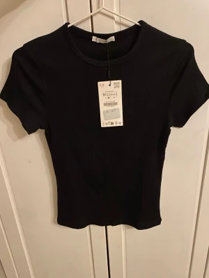 Svart ribbad topp från Zara - En svart ribbad topp från Zara med rund halsringning och korta ärmar. Toppen har en tight passform och är tillverkad i ett mjukt, stretchigt material som sitter snyggt på kroppen. Aldrig använd och prislappen kvar.