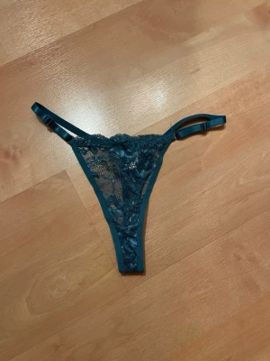 Mörkgröna spets-trosor med smala band XL - Snygga mörkgröna trosor i stringmodell med tunna, justerbara band i sidan. Trosorna är gjorda i genomskinlig spets med blommigt mönster och har en feminin look. Perfekta för dig som gillar detaljer och en trendig stil. Färg petroleum blå.