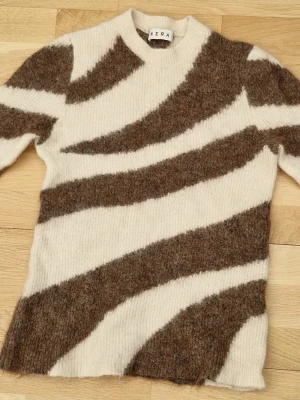 Stickad tröja med zebramönster från Wera - Säljer en stickad tröja från Wera med zebrainspirerat mönster i brunt och offwhite. Tröjan har rund hals, lång ärm och är gjord i ett mjukt, lurvigt material som känns riktigt cozy. Perfekt för dig som vill sticka ut lite extra. Storleken är större än vad som sägs (enligt mig). Denna var för stor för mig och jag brukar ha XS/XXS.