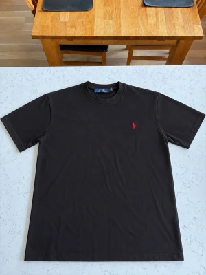 Svart t-shirt från Polo Ralph Lauren - Svart t-shirt från Polo Ralph Lauren med klassisk passform och korta ärmar. T-shirten har en liten röd broderad logga på bröstet och är tillverkad i mjuk bomull. Perfekt för en clean och stilren look.