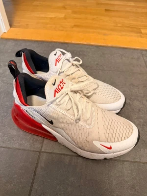 Nike Air Max 270 vita och röda sneakers - Nike Air Max 270 sneakers i vitt med röda och svarta detaljer. Ovandel i mesh för bra ventilation, stor synlig Air Max-dämpning i hälen och klassisk Nike-logga på sidan. Snygg och sportig design som sticker ut med den röda häldelen.