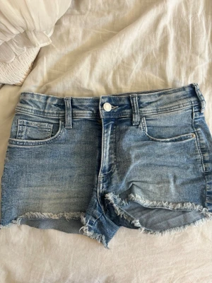 Blå jeansshorts med fjärilsbroderi - Säljer ett par ljusblå jeansshorts med råa kanter och femficksmodell. På ena bakfickan finns ett snyggt broderat fjärilsmotiv som ger shortsen en unik touch. Perfekta för varma dagar och enkel att matcha med olika toppar.