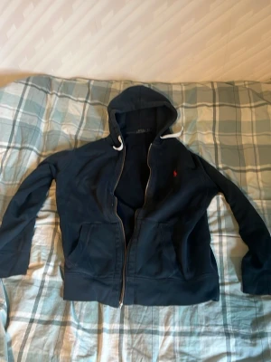 Marinblå hoodie från Polo Ralph Lauren - Snygg marinblå hoodie från Polo Ralph Lauren med dragkedja, två fickor framtill och vit dragsko i huvan. Klassisk röd logga broderad på bröstet. Tillverkad i mjukt bomullsmaterial som är skönt att ha på sig. Perfekt för en avslappnad och stilren look.