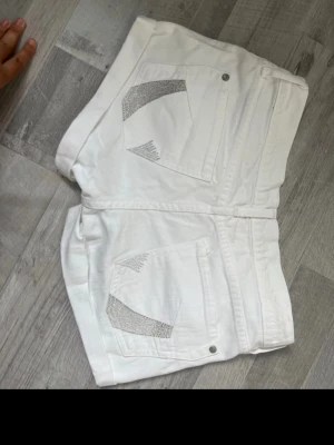 Vita höga shorts med silverdetaljer - Säljer ett par vita shorts med hög midja och snygga silverfärgade detaljer på bakfickorna. Klassisk femficksmodell med knapp och dragkedja framtill. Perfekta till sommaren och riktigt fräscha till alla typer av toppar.