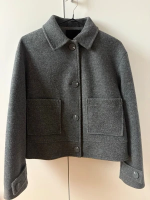 Grå kappa från 157, XS - Stilren grå kappa från 157 i ullmix med stora framfickor och klassisk krage. Modellen är croppad och har stora knappar framtill samt vid ärmsluten. Perfekt för dig som gillar minimalistisk och trendig look.