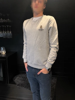 Grå sweatshirt från Morris - Snygg grå sweatshirt från Morris med broderad logga på bröstet. Tröjan har rund halsringning, långa ärmar och ribbade muddar vid ärmslut och nederkant. Perfekt för en avslappnad och stilren look.
