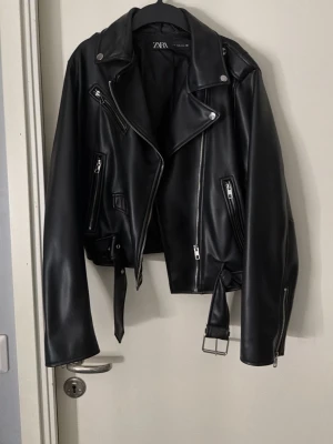 Svart skinnjacka från ZARA - Snygg svart skinnjacka från ZARA med klassisk bikerstil. Jackan har flera dragkedjeförsedda fickor, bred krage med tryckknappar och ett bälte i midjan. Silverfärgade detaljer och raka ärmar med dragkedjor ger en cool look.