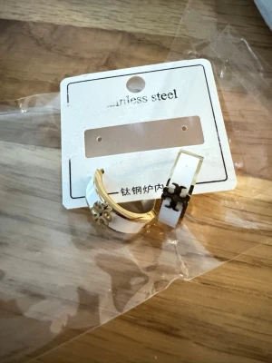 Vita örhängen i rostfritt stål Tory Burch inspiration - Snygga örhängen i vit och guld färg med dekorativa detaljer i metall. Tillverkade i rostfritt stål och har en modern, stilren design med inslag av geometriska former. Perfekta för att lyfta din outfit med en trendig touch. Endast inspiration ej äkta