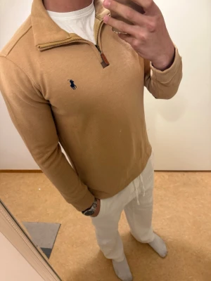 Beige stickad tröja med halv dragkedja - Snygg beige stickad tröja från Polo Ralph Lauren med halv dragkedja och klassisk blå logga på bröstet. Tröjan har ribbade muddar och hög krage, perfekt för lager på lager. Materialet känns mjukt och bekvämt.