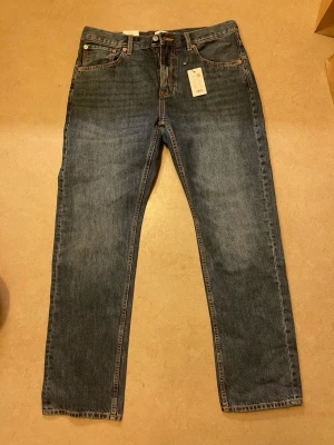 Blå raka jeans med hög midja - Klassiska blå jeans med rak passform och hög midja. Jeansen har fem fickor, bälteshällor och stängs med knapp och dragkedja. Materialet är robust denim i 100% bomull. Perfekta för en avslappnad och tidlös stil.
