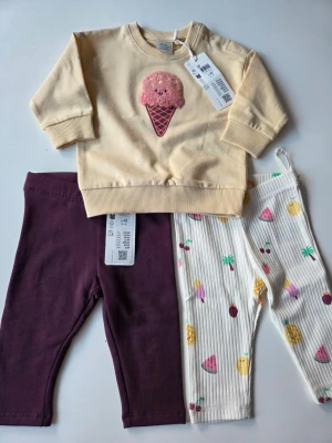 Sött set med glass-tröja och 2 leggings strl 62 - Set med ljusgul sweatshirt med broderad glass i rosa och brunt framtill, samt två par leggings: ett par i vinrött med lite foder i och ett par vita med färgglada fruktmotiv. Perfekt för dig som gillar lekfulla och färgglada plagg. Mjukt material och bekväm passform.