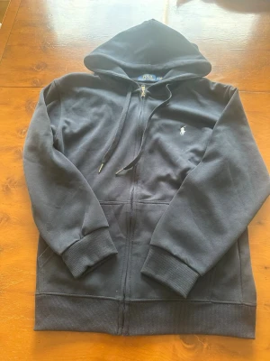 Mörkblå Ralph lauren zip hoodie - Säljer en svart hoodie med dragkedja från Polo Ralph Lauren. Klassisk design med huva och dragsnören, ribbade muddar och fickor framtill. Liten vit logga broderad på bröstet. Perfekt för en chill och stilren look.