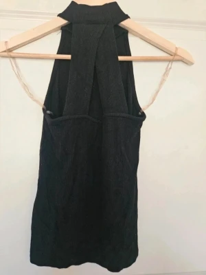 Svart ribbad polotopp Amisu S - Snygg svart ribbad polotopp med hög hals och ärmlös design. Toppen har breda axelband som korsar i ryggen för en cool detalj. Perfekt att styla med jeans eller kjol för en trendig look.  E4