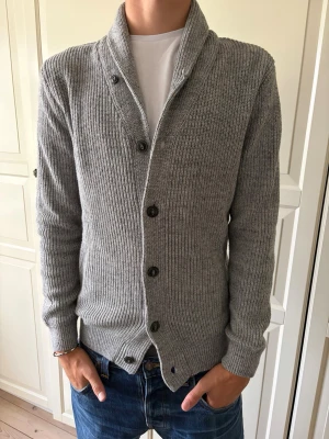 Grå ribbstickad kofta med sjalkrage - Snygg grå ribbstickad kofta med sjalkrage och knäppning framtill. Koftan har långa ärmar och en klassisk passform som passar till jeans eller chinos. Perfekt för lager-på-lager och enkel att styla till olika outfits. Tröjan är i storlek xs men passar som S. Hör av er vid frågor och priset är inte hugget i sten!