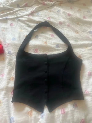 Svart halterneck väst från Gina Tricot - Snygg svart väst från Gina Tricot i halterneck-modell med knappar framtill. Västen har en figurnära passform och spetsig nederkant, perfekt för att lyfta vilken outfit som helst.