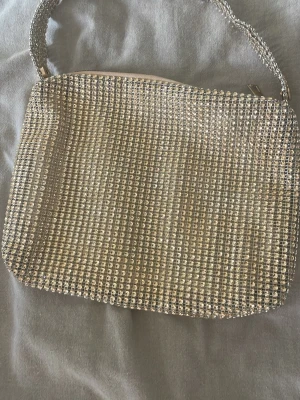 Glittrig handväska med strass - Säljer en superglittrig handväska täckt med silvriga strass-stenar. Väskan har en mjuk, rektangulär form och ett matchande handtag med dragkedja upptill. Insidan är ljus och rymlig, perfekt för det viktigaste. Perfekt accessoar för att lyfta vilken outfit som helst!