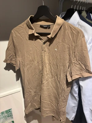 Beige pikétröja från J.Lindeberg - Snygg beige pikétröja från J.Lindeberg med klassisk krage och två knappar framtill. Tröjan har korta ärmar och är tillverkad i ett mjukt bomullsmaterial. Liten vit logga broderad på bröstet för en clean look.