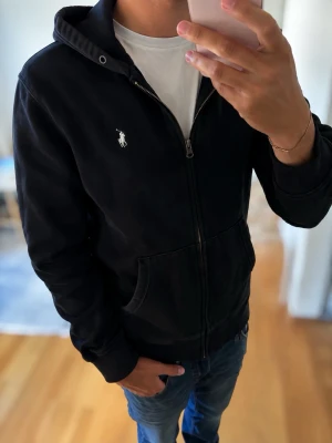 Svart Ralph Lauren Zip - Tröja - En skön svart Ralph Lauren Tröja med vit emblem. Passar till dig som vill hålla värmen utan att förlora stilen. Perfekt nu till Höst och Skolstart.