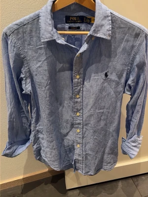 Blå linneskjorta från Polo Ralph Lauren - Snygg ljusblå skjorta i linne från Polo Ralph Lauren med klassisk krage, knappar framtill och broderad logga på bröstet. Skjortan har lång ärm och en avslappnad men ändå skräddarsydd passform. Perfekt för en stilren look.