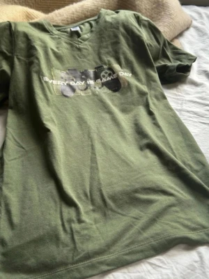 Olivgrön t-shirt med tryck - Säljer en olivgrön t-shirt med coolt tryck på bröstet där det står 'EVERY DAY IS GAME DAY' och en grafisk design med speltema. T-shirten har rund hals och korta ärmar. Perfekt för dig som gillar gaming och vill ha en avslappnad stil.