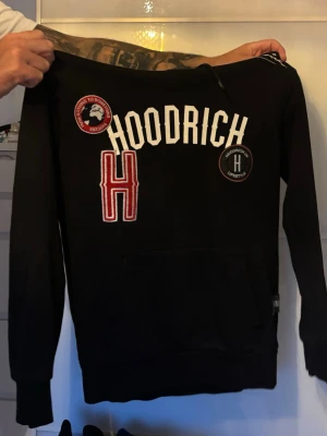 Svart hoodie från Hoodrich - Svart hoodie med luva från Hoodrich med vita och röda broderade detaljer och patches på bröstet och även en ficka. Knappa använd och ser ut som ny 