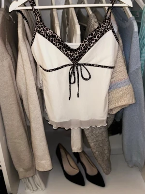 Vit topp med leoparddetaljer - Snygg vit topp med tunna axelband och leopardmönstrade detaljer längs urringningen och under bysten. Dekorativ knytning framtill och lätt vågig kant nertill. Perfekt för att addera lite edge till din outfit.
