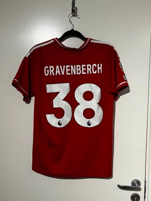 Liverpool matchtröja Gravenberch 38 - Röd Liverpool matchtröja med vita detaljer och tryck 'Gravenberch 38' på ryggen. Framtill finns Liverpool FC-logga, Adidas-logga och sponsortryck. Korta ärmar och sportig passform, perfekt för fotbollsträning eller match.