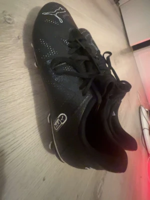 Svarta fotbollsskor från Puma - Snygga svarta fotbollsskor från Puma med vita detaljer och klassisk logga på tån och innersulan. Skorna har snörning och dobbar för bra grepp på planen. Tillverkade i syntetmaterial som ger en lätt och smidig känsla när du spelar.
