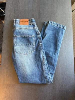Blåa slim fit jeans från Sam’s Jeans - Snygga blåa slim fit jeans från Sam’s Jeans med klassisk femficksdesign och läderpatch bak i midjan. Jeansen har en tvättad look med ljusare partier och raka ben. Perfekta för en avslappnad och trendig stil.